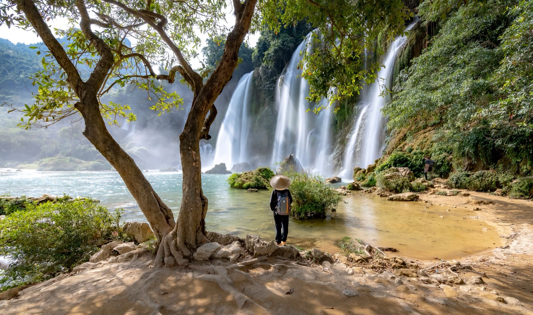 Cao Bang — A lone traveler stands by the majestic Ban Gioc waterfall, surrounded by lush gr