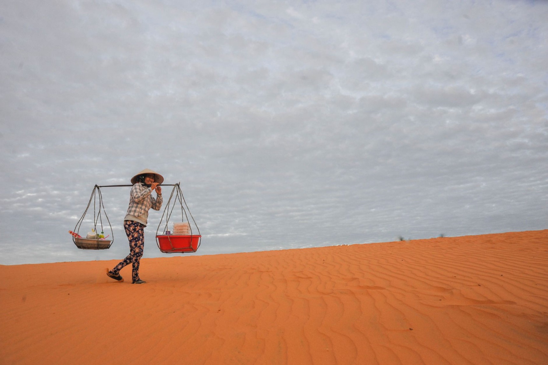 Mui Ne — Woman carrying goods with a conical hat on a desert sand dune, symbolizing resil