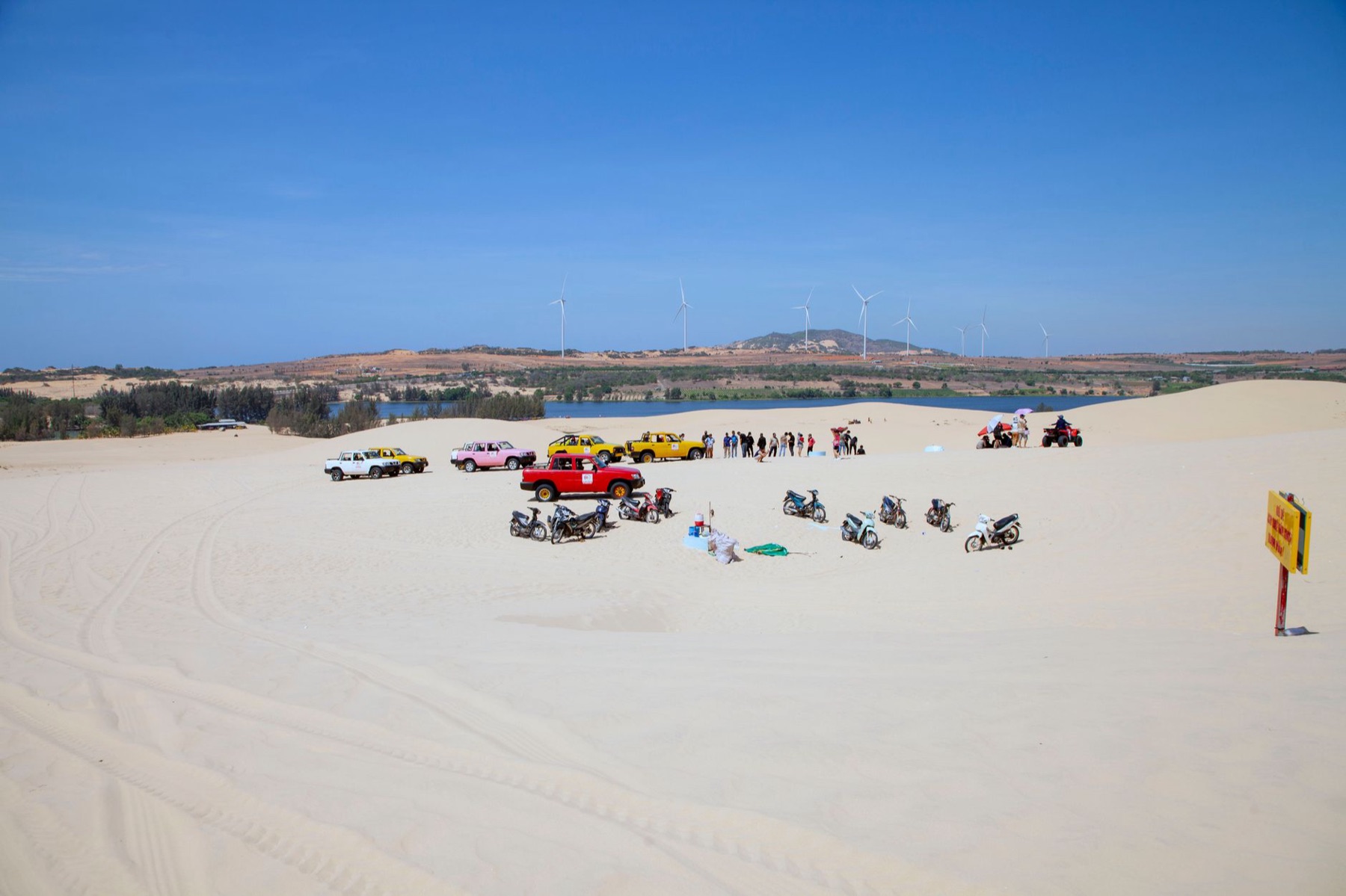 Mui Ne — Tourists enjoy a sunny dune landscape with colorful vehicles and distant wind tu